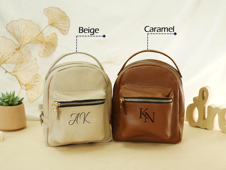 May include: Two small pebbled leather backpacks with top handles and front zippered pockets. The beige backpack features the monogram A.K., while the caramel backpack displays the monogram K.N. Both bags have gold-toned hardware and zippers.