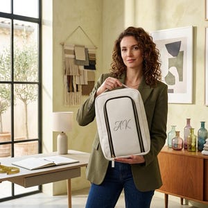 May include: A rectangular off-white travel organizer bag featuring a black zippered front pocket and the embroidered monogram initials AK in a cursive script. The bag includes a top handle and a structured design with black piping along the edges.