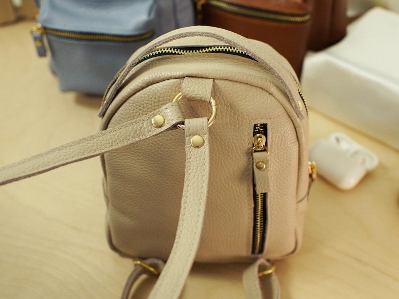 May include: A beige pebbled leather mini backpack featuring gold-tone metal hardware, a vertical zippered pocket on the back, and adjustable shoulder straps attached to a central gold ring with metal rivets.