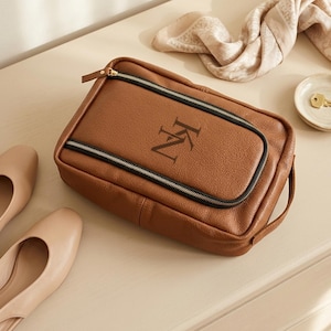 May include: A rectangular brown leather toiletry bag with a pebbled texture, featuring a black zippered front pocket and a black zipper closure. The front of the bag is embossed with the letters KN in a serif font. A side handle is attached.