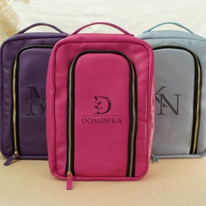 May include: Three rectangular leather bags in purple, hot pink, and light blue. Each bag features a top handle, a vertical zippered front compartment, and a personalized monogram. The center pink bag is clearly marked with the name DOMINIKA.