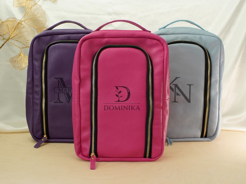 May include: Three rectangular leather bags in purple, hot pink, and light blue. Each bag features a top handle, a vertical zippered front compartment, and a personalized monogram. The center pink bag is clearly marked with the name DOMINIKA.