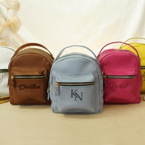May include: Five small backpacks in white, brown, light blue, hot pink, and yellow, each featuring a top handle, a front zippered pocket, and personalized black lettering. The bags have a textured surface and gold-toned metal zipper hardware.