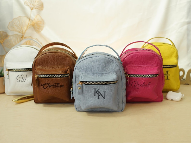 May include: Five small backpacks in white, brown, light blue, hot pink, and yellow, each featuring a top handle, a front zippered pocket, and personalized black lettering. The bags have a textured surface and gold-toned metal zipper hardware.