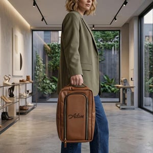 May include: A rectangular brown leather-like travel bag with a top handle and a vertical zippered front pocket. The bag features the name Adam embroidered in black script on the front panel. The design includes black trim along the zippers.