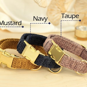 May include: Three dog collars in mustard, navy, and taupe. Each collar has a gold-colored metal buckle and a matching D-ring. The collars are made of a textured fabric and are displayed on a light-colored surface.