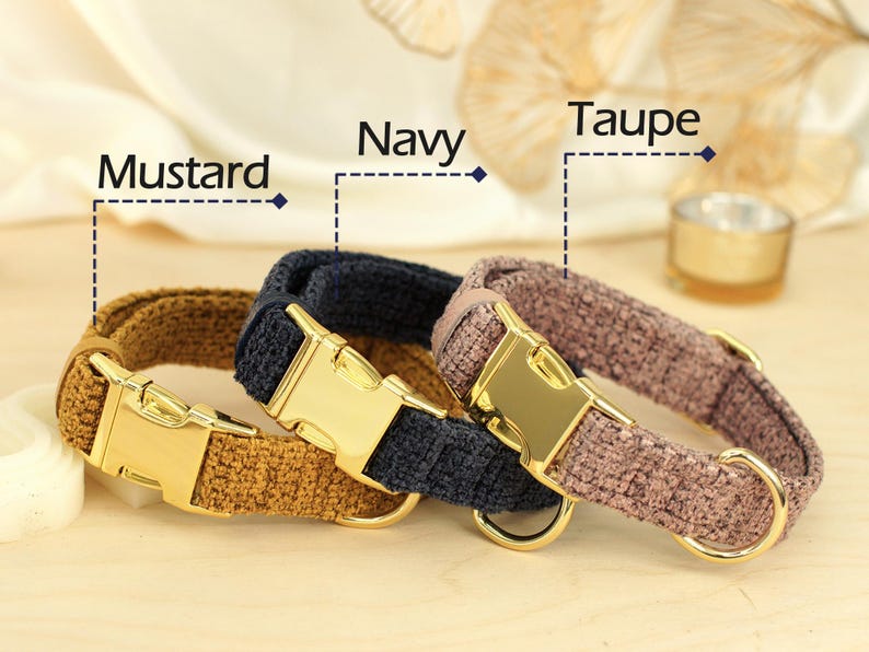 May include: Three dog collars in mustard, navy, and taupe. Each collar has a gold-colored metal buckle and a matching D-ring. The collars are made of a textured fabric and are displayed on a light-colored surface.