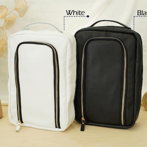 May include: Two rectangular travel organizers with a textured finish, one in white and one in black. Each bag features a top handle, a vertical U-shaped front zipper closure with gold-toned hardware, and the labels White and Black above them.