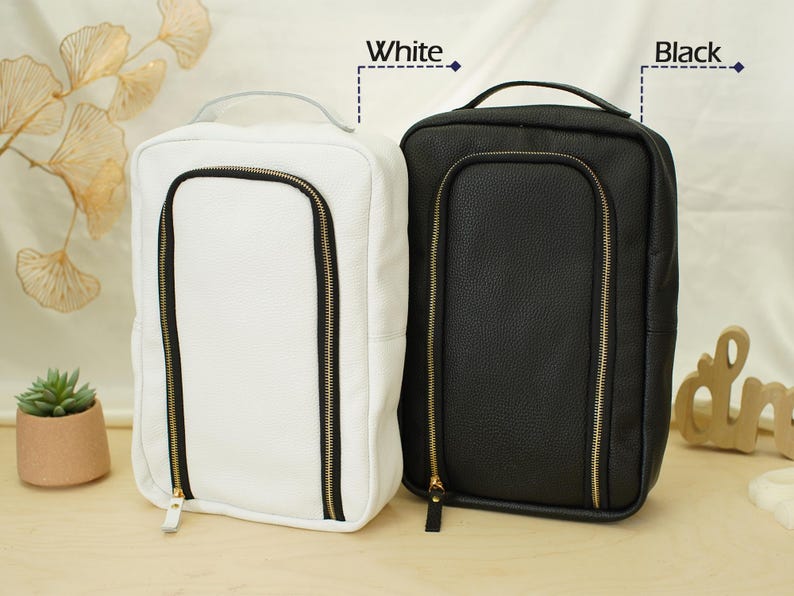 May include: Two rectangular travel organizers with a textured finish, one in white and one in black. Each bag features a top handle, a vertical U-shaped front zipper closure with gold-toned hardware, and the labels White and Black above them.