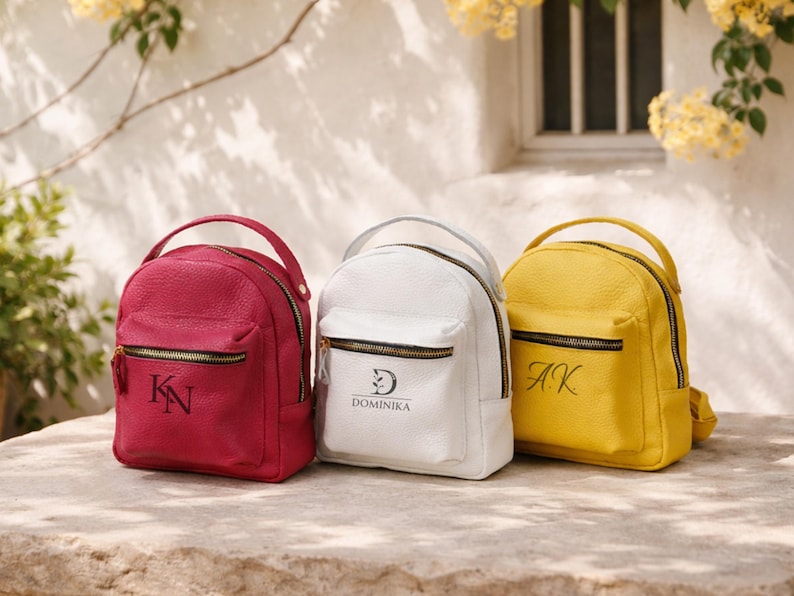 May include: Three small backpacks in magenta, white, and yellow with a pebbled texture. Each bag features a top handle, a front zippered pocket, and black metal hardware. The magenta bag has the letters KN, the white bag says DOMINIKA, and the yellow bag has AK.