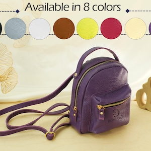 May include: A purple mini backpack with a top handle, adjustable shoulder straps, and a front zippered pocket. The text "Available in 8 colors" appears above a row of eight circular color swatches including black, blue, white, brown, yellow, pink, cream, and purple.