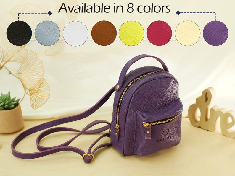 May include: A purple mini backpack with a top handle, adjustable shoulder straps, and a front zippered pocket. The text "Available in 8 colors" appears above a row of eight circular color swatches including black, blue, white, brown, yellow, pink, cream, and purple.