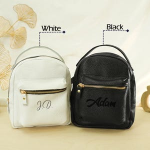 May include: Two small backpacks with a pebbled texture, one white and one black. Each bag features a top handle, a front zippered pocket with gold-tone hardware, and personalized text. The white bag is embroidered with J.D. and the black bag with Adam.