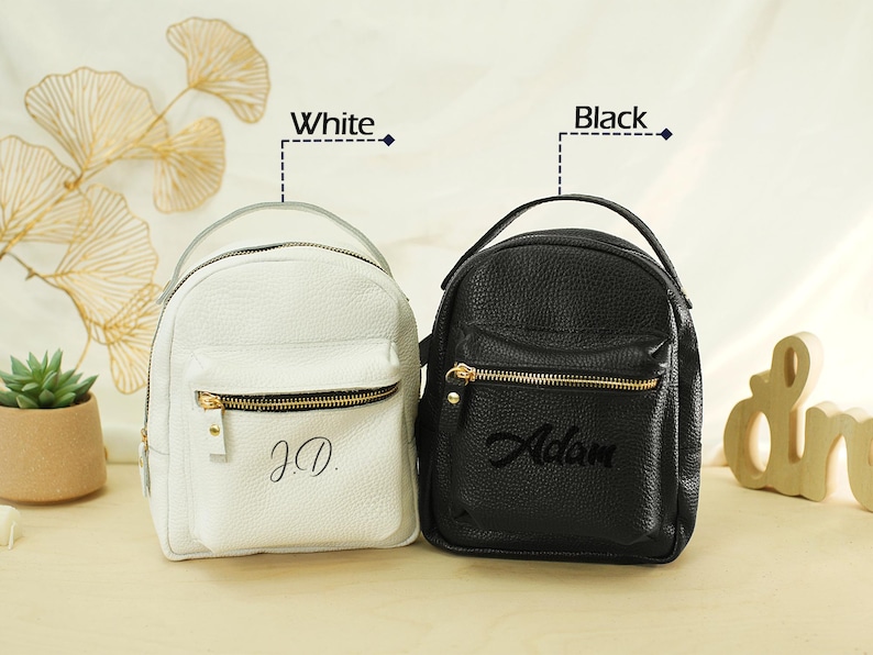May include: Two small backpacks with a pebbled texture, one white and one black. Each bag features a top handle, a front zippered pocket with gold-tone hardware, and personalized text. The white bag is embroidered with J.D. and the black bag with Adam.