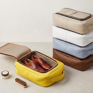 May include: A stack of five rectangular zippered shoe bags in beige, white, light blue, brown, and yellow. The yellow bag is open, containing a pair of brown leather oxford shoes. A small round container and a wooden shoe brush are placed nearby.