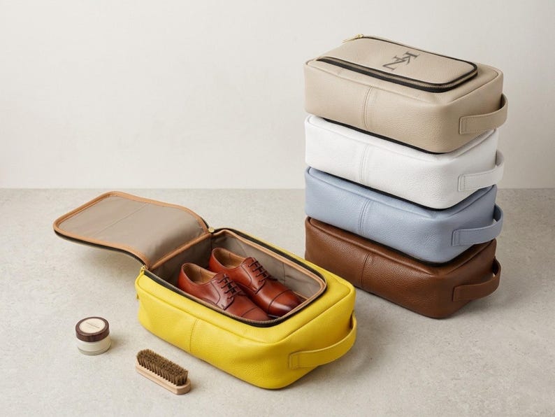 May include: A stack of five rectangular zippered shoe bags in beige, white, light blue, brown, and yellow. The yellow bag is open, containing a pair of brown leather oxford shoes. A small round container and a wooden shoe brush are placed nearby.