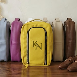 May include: A collection of rectangular leather shoe bags in various colors including light blue, purple, yellow, beige, and brown. The yellow bag features a black zippered front compartment with the embroidered monogram letters KN in dark green.