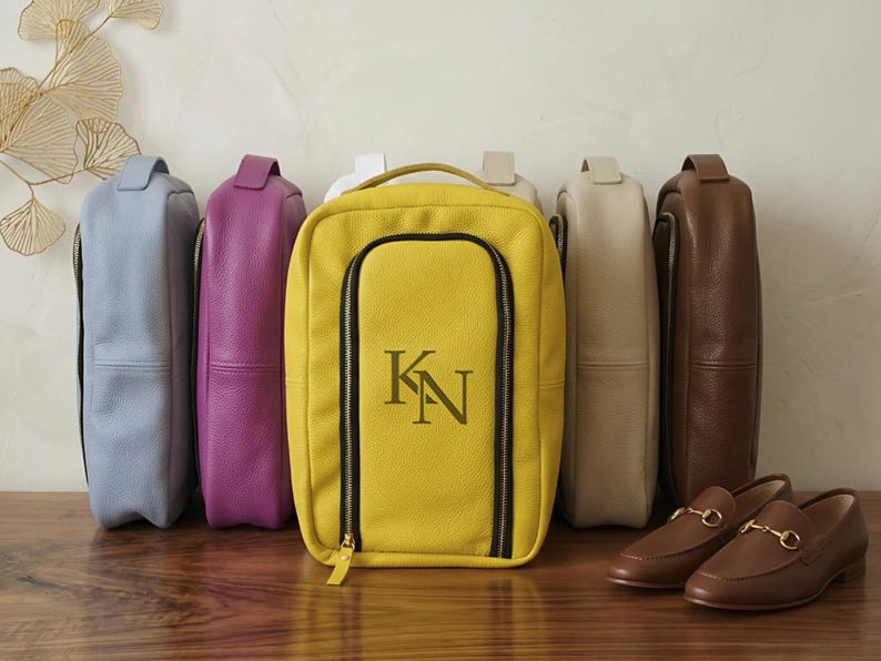May include: A collection of rectangular leather shoe bags in various colors including light blue, purple, yellow, beige, and brown. The yellow bag features a black zippered front compartment with the embroidered monogram letters KN in dark green.