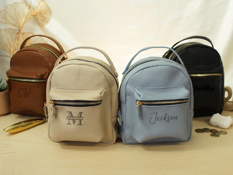 May include: Four small, pebbled leather mini backpacks in brown, beige, light blue, and black. Each bag features a top handle, a front zippered pocket, and gold-tone hardware. The beige bag is monogrammed with 'MARLIN', and the blue bag with 'Jackson'.
