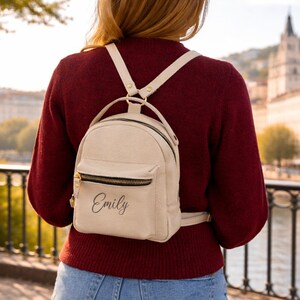 May include: A small, cream-colored backpack featuring a front zippered pocket with the name Emily printed in black script. The bag includes a top handle and adjustable shoulder straps with gold-toned metal hardware and black zipper detailing.