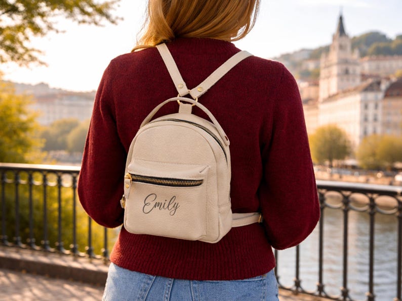 May include: A small, cream-colored backpack featuring a front zippered pocket with the name Emily printed in black script. The bag includes a top handle and adjustable shoulder straps with gold-toned metal hardware and black zipper detailing.