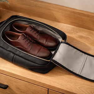 May include: A black textured leather shoe bag with a gold-toned zipper, partially open to reveal a pair of dark brown leather oxford shoes with laces inside. The interior of the bag is lined with a light gray fabric, and the exterior features a handle.