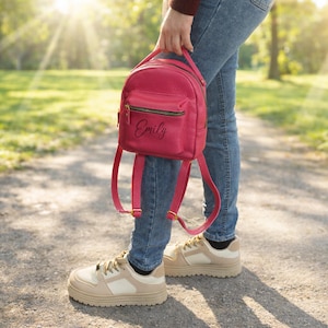May include: A small, vibrant pink backpack featuring a textured surface, a top handle, adjustable shoulder straps, and a front zippered pocket. The name Emily is printed in black script on the front pocket, which also includes gold-toned hardware.