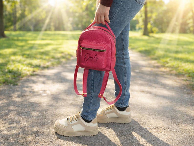 May include: A small, vibrant pink backpack featuring a textured surface, a top handle, adjustable shoulder straps, and a front zippered pocket. The name Emily is printed in black script on the front pocket, which also includes gold-toned hardware.