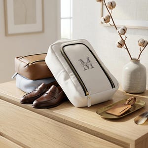 May include: A white leather toiletry bag with a black zipper and the monogrammed name Martin, accompanied by a tan leather bag, a light blue bag, a pair of brown leather loafers, a tan leather luggage tag on a gold tray, and a shoe horn.