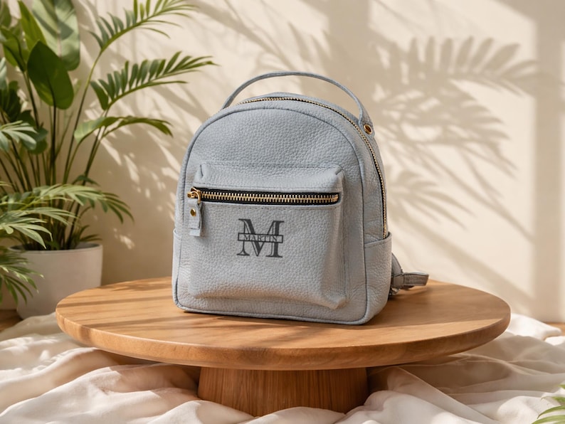 May include: A light blue pebble-textured mini backpack featuring a top handle, a front zippered pocket, and a dark grey monogram logo with the letter M and the name MARTIN printed in the center. The bag has gold-toned metal zipper hardware.