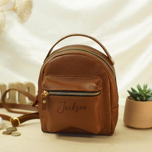 May include: A small, brown, pebbled-texture backpack with a top handle and a front zippered pocket. The name Jackson is printed in black script on the front pocket. The bag features gold-toned metal zippers and hardware, along with adjustable straps.