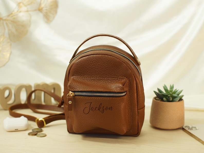 May include: A small, brown, pebbled-texture backpack with a top handle and a front zippered pocket. The name Jackson is printed in black script on the front pocket. The bag features gold-toned metal zippers and hardware, along with adjustable straps.