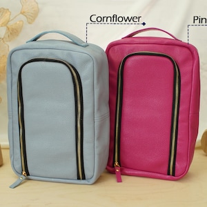 May include: Two rectangular travel cases with top handles and U-shaped front zippers. One case is cornflower blue and the other is pink. Both feature black zipper tape with gold-toned hardware and a textured exterior finish.