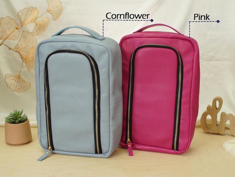 May include: Two rectangular travel cases with top handles and U-shaped front zippers. One case is cornflower blue and the other is pink. Both feature black zipper tape with gold-toned hardware and a textured exterior finish.