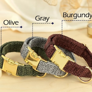 May include: Three dog collars in olive, gray, and burgundy. Each collar has a gold-colored metal buckle and a matching D-ring. The collars are made of a textured fabric and are displayed on a light-colored wooden surface.