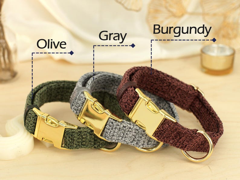 May include: Three dog collars in olive, gray, and burgundy. Each collar has a gold-colored metal buckle and a matching D-ring. The collars are made of a textured fabric and are displayed on a light-colored wooden surface.