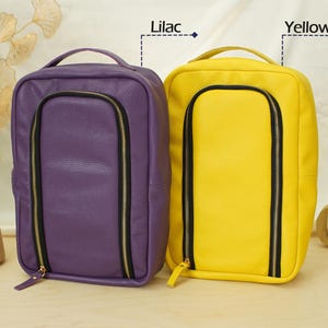 May include: Two rectangular bags with a textured finish, one in lilac and one in yellow. Each bag features a top handle, a vertical U-shaped front zipper with black trim and gold hardware, and a clean, structured design for organized storage.