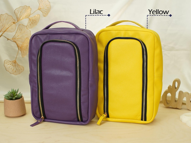 May include: Two rectangular bags with a textured finish, one in lilac and one in yellow. Each bag features a top handle, a vertical U-shaped front zipper with black trim and gold hardware, and a clean, structured design for organized storage.