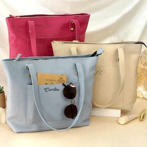 May include: Three leather tote bags in pink, light blue, and cream. The light blue bag has a front pocket with a notebook, sunglasses, and the name "Caroline" embroidered. The cream bag has initials embroidered. All bags have top handles.