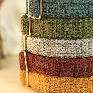 May include: A stack of five dog collars in various colors: teal, olive green, gray, rust, and mustard yellow. Each collar has a textured, plush appearance and is adorned with gold-colored metal hardware.