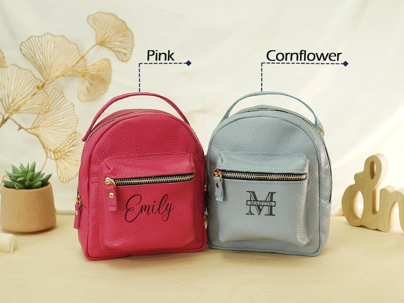 May include: Two small backpacks with top handles and front zippered pockets. The left backpack is pink with the name Emily printed in black. The right backpack is cornflower blue with the name Martin and a letter M monogram printed in black.