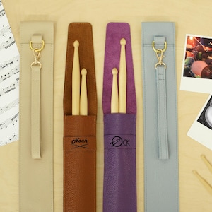 May include: Four leather drumstick cases in beige, brown, purple, and blue-grey, each with a gold-colored clasp and strap. The brown and purple cases have personalized text. Two drumsticks and two polaroid photos of a drum set are also visible.