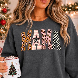 May include: A dark heather gray crewneck sweatshirt featuring the word MAMA printed in large block letters. The letters have a mixed pattern design including leopard print, pink and white horizontal stripes, and black and white zebra stripes.