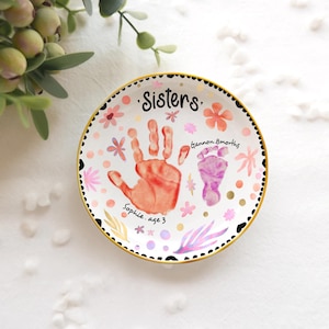 Op de afbeelding: Een witte keramische plaat met een gouden rand, met een hand- en voetafdrukontwerp. De plaat is versierd met bloemaccenten en het woord "Sisters". De handafdruk is oranje en de voetafdruk is paars.