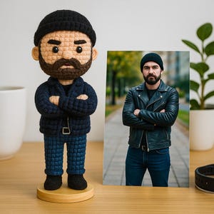 Può includere: Bambola all'uncinetto fatta a mano di un uomo con la barba, con berretto nero, giacca blu e jeans. La bambola è su una base di legno. Accanto, una foto incorniciata di un uomo con giacca di pelle nera e jeans.