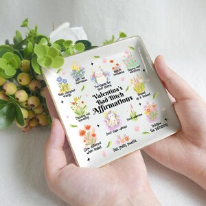 Könnte beinhalten: Ein quadratisches, cremefarbenes Keramik-Schmucktablett mit goldenem Rand. Das Tablett zeigt den Text "Valentina's Bad Bitch Affirmations" und verschiedene Blumenillustrationen mit positiven Affirmationen. Das Tablett wird in den Händen einer Person gehalten.
