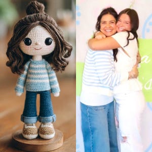 May include: A handmade crochet doll with brown hair styled in a bun and wavy locks. The doll wears a blue and white striped sweater, blue jeans, and tan shoes. The doll is standing on a wooden base. Two women are hugging in the background.