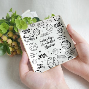 Könnte beinhalten: Ein quadratisches, weißes Keramiktablett mit goldenem Rand, das Volleyball-Illustrationen und motivierende Sätze in Schwarz zeigt. Der Text enthält "Hailey's Serve Like a Goddess Affirmations". Das Tablett wird von einer Person gehalten.