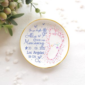 Op de afbeelding: Een witte keramische plaat met een gouden rand, versierd met een babyvoetprintontwerp in roze en paarse stippen. De plaat bevat de tekst "I love you daughter", een naam, datum, tijd, locatie en een afmeting van 59 cm.