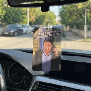 May include: Rectangular car air freshener with a printed image of a man on a phone. The air freshener has text in Turkish. The image is hanging from a car's rearview mirror, with the dashboard visible.